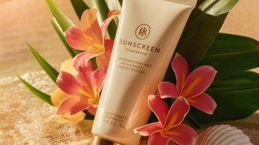 Luxury sunscreen tube with cream and gold packaging on sandy beach surrounded by seashells and tropical flowers under warm sunlight.