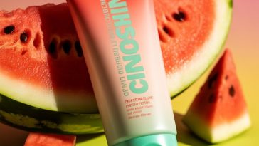 A vibrant watermelon-themed sunscreen tube with coral-red to light-green packaging surrounded by fresh watermelon slices on a pink-to-green gradient background.